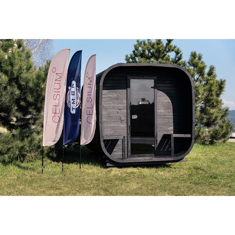 Frontansicht der schwarzen Celsium Ula Large Sauna im Freien neben drei Event-Fahnen vor grüner Kulisse