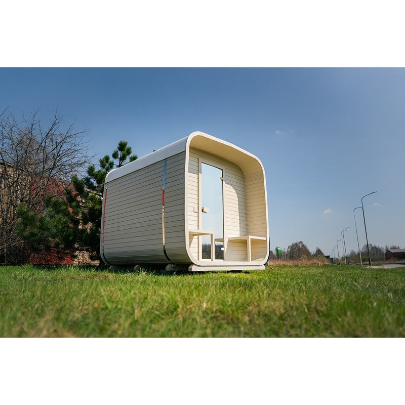 Außenansicht der weißen Celsium Ula Large Sauna mit überdachtem Vorraum und Glastür auf einer grünen Wiese bei klarem Himmel