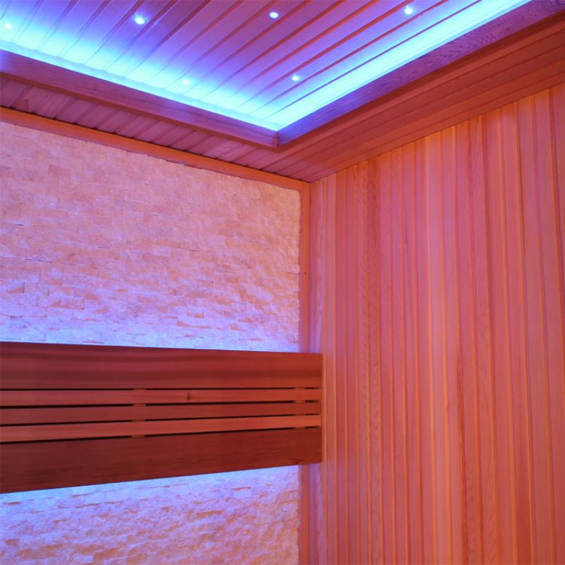 Modernes Saunakabineninterior mit roten Zedernholzwänden, breiter Liege mit Rückenlehne, strukturierter Rückwand und LED-Deckenbeleuchtung in Blau- und Orangetönen
