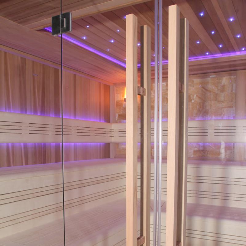 Moderne Sauna mit roter Zeder, Glastür und atmosphärischer Beleuchtung in Blau- und Orangetönen, Holzbänke und Deckenleuchten sichtbar.