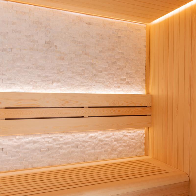 Innenansicht einer modernen Sauna mit Pinieholzverkleidung, Saunaliegen aus hellem Holz mit Rückenlehne und beleuchteter Natursteinkachel-Wand
