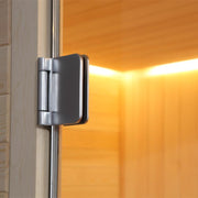 Detailaufnahme des verchromten Türscharniers einer Sauna mit Pinienholzrahmen und orangegelbem Glasbereich im Hintergrund