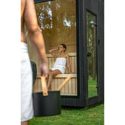 Frau entspannt in der Celsium Jonas Small Glassauna – stilvolle Gartensauna aus Holz mit großem Glasfenster und natürlichem Lichteinfall.