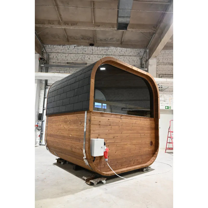 Modular aufgebaute Fasssauna Hekla Cube mit abgerundetem Dach, Holzverkleidung in Natur und schwarzem Dachschindeln, großem Panoramafenster, in Werkshalle aufgestellt
