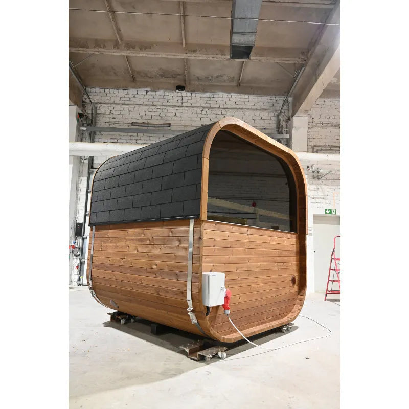 Hekla Cube Fass-Sauna aus Holz mit schwarzem Dach in einer Lagerhalle, Vorderansicht mit Glastür und Holzverkleidung