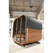 Hekla Cube Sauna mit abgerundeter Holzform, schwarzem Bitumen-Dachschindeln-Dach und großer Glastür in Bauhalle stehend