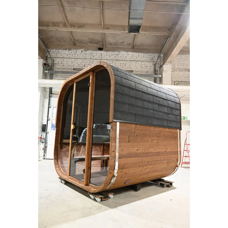 Hekla Cube Sauna mit abgerundeter Holzform, schwarzem Bitumen-Dachschindeln-Dach und großer Glastür in Bauhalle stehend