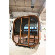 Hekla Cube Sauna aus dunklem Holz mit großen Glastüren und Holzbank in einer Produktionshalle