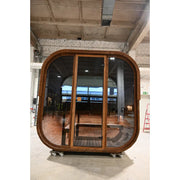 Moderne Sauna Hekla Cube mit abgerundeter Holzkonstruktion, großen Glastüren und Holzboden in industrieller Ausstellungshalle