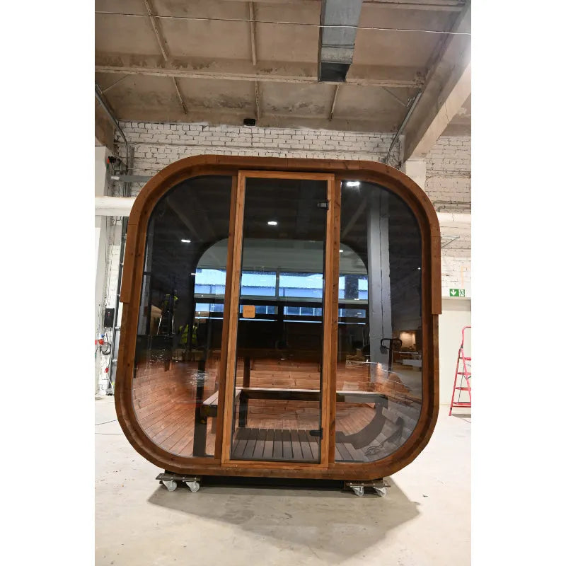 Moderne Sauna Hekla Cube mit abgerundeter Holzkonstruktion, großen Glastüren und Holzboden in industrieller Ausstellungshalle