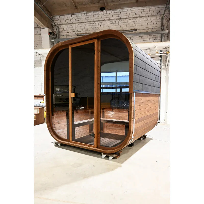 Moderne Fasssauna Hekla Cube mit abgerundeter Holzfront, großen Glastüren und Holzbank im Innenraum, in einer Fabrikhalle aufgestellt