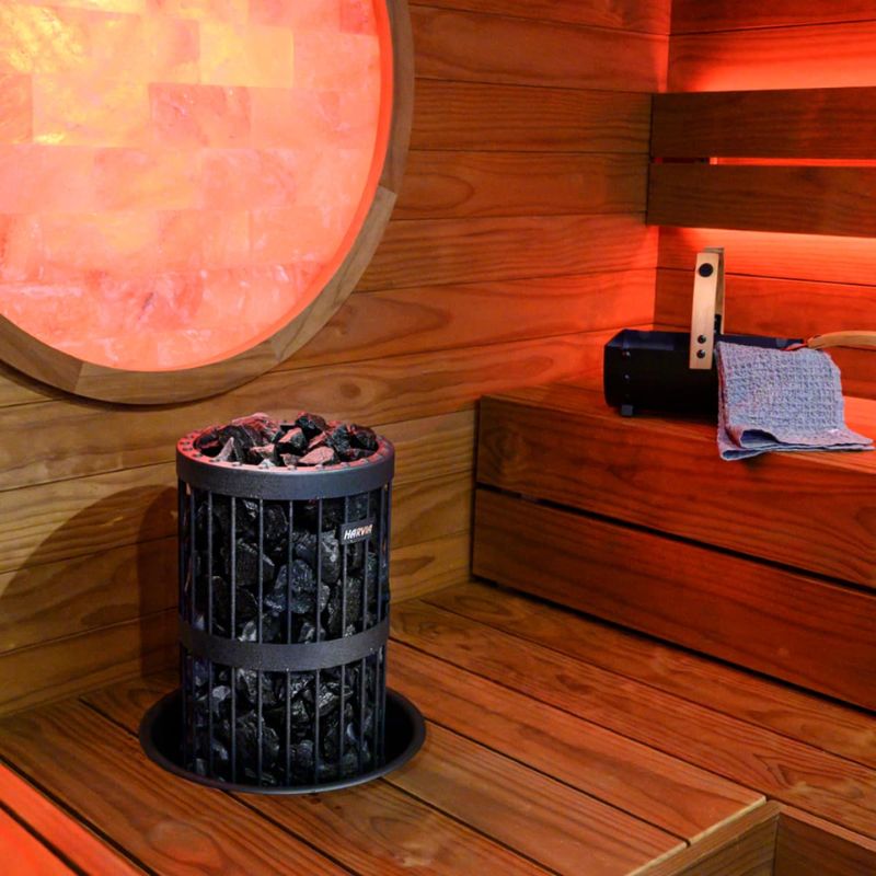 Harvia Legend Home Saunaofen mit glühenden Steinen in moderner Holzsauna, daneben Infrarotwärmepaneel und Handtuch auf Holzbank