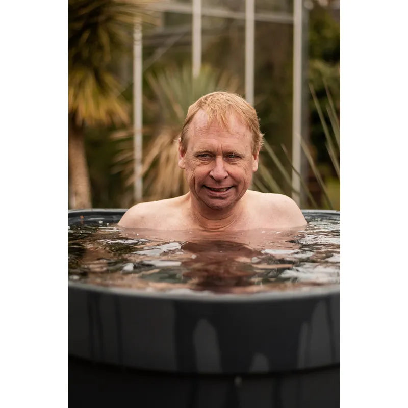 Mann badet lächelnd im Passion Revive Pro Eisbad-Kältebecken mit tropischem Glashaus im Hintergrund