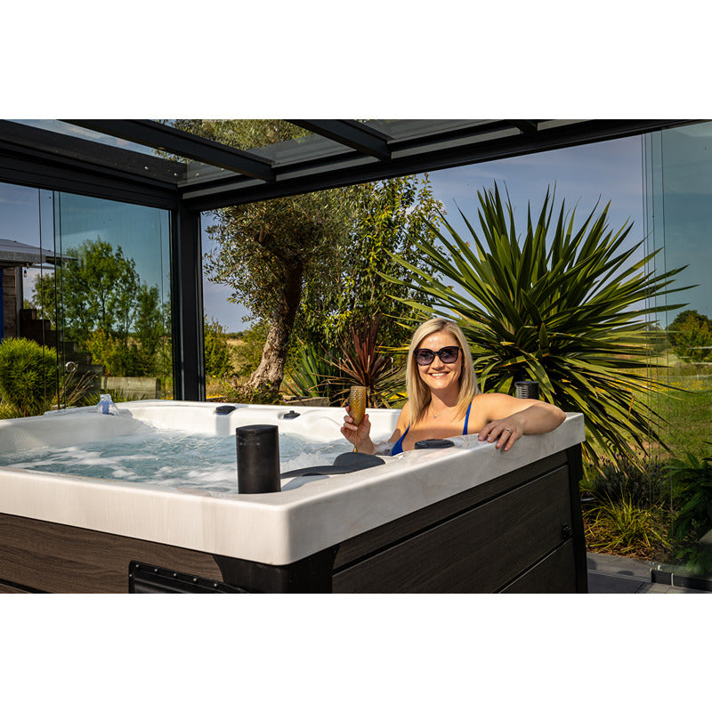Frau entspannt in einem weißen Platinum Spas Topaz Outdoor Whirlpool unter Glasschiebedach mit Blick auf grüne Gartenlandschaft und Palmen.
