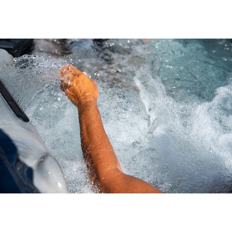 Nahaufnahme: Hand berührt den Wasserfallstrahl im Platinum Spas Topaz Outdoor Whirlpool – Premium. Sprudelndes Wasser und Sonnenlicht.