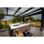 Frau entspannt sich im Platinum Spas Topaz Whirlpool unter moderner schwarzer Glasüberdachung mit Blick auf grüne Gartenlandschaft und Palmen.