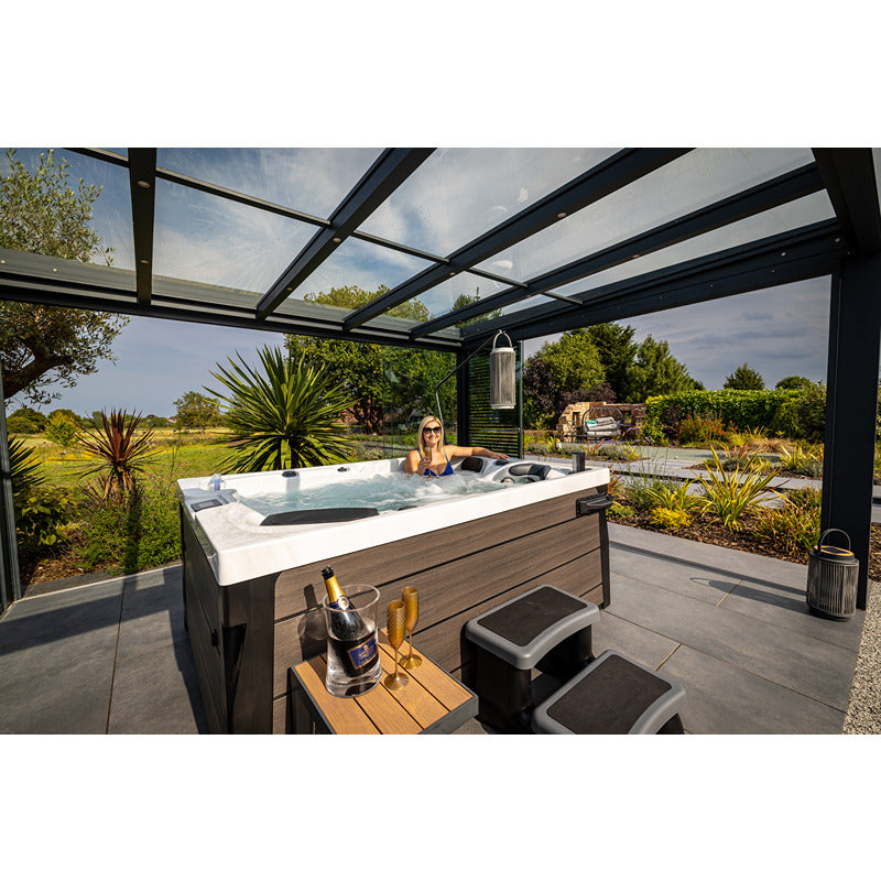 Frau entspannt sich im Platinum Spas Topaz Whirlpool unter moderner schwarzer Glasüberdachung mit Blick auf grüne Gartenlandschaft und Palmen.