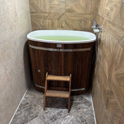 Eisbad aus dunklem Thermoholz mit weißem Innenrand, gefüllt mit grünem Wasser, in moderner Badezimmerecke mit geometrischen Holzfliesen und Holztreppe