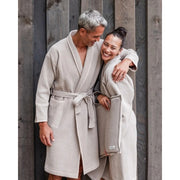 Ein Paar in Badesofa Bademänteln aus Bio-Baumwolle lehnt lachend aneinander vor einer Holzfassade.