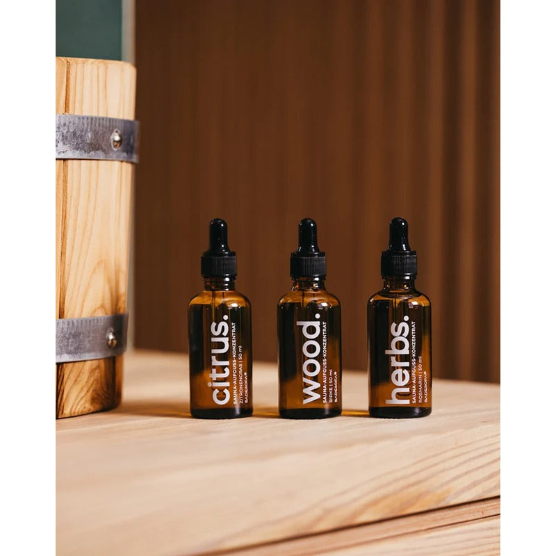 Drei Fläschchen des Badesofa Saunaaufguss-Sets mit Pipetten und den Duftnoten Citrus, Wood und Herbs auf einer Holzbank