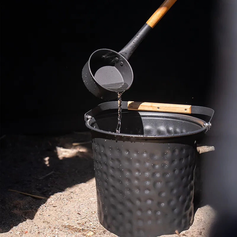 Bast Näver Deluxe Zeltsauna Zubehör: Wasserschöpfer füllt schwarzen Saunaeimer mit Holzgriffen