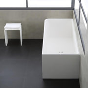 Moderne, freistehende Badewanne aus weißem Mineralguss mit rechteckiger Form, daneben ein kleiner weißer Ablagehocker mit Handtuch in minimalistischem Badezimmer