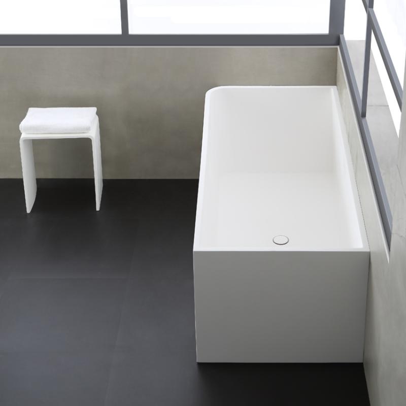 Moderne, freistehende Badewanne aus weißem Mineralguss mit rechteckiger Form, daneben ein kleiner weißer Ablagehocker mit Handtuch in minimalistischem Badezimmer