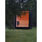 Moderne Außensauna Jonas Small im Naturumfeld, durch Nadelbaum teils verdeckt, mit stimmungsvoll beleuchtetem Innenraum.