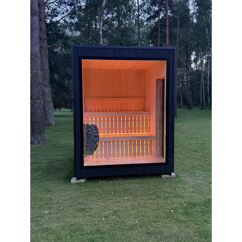 Freistehende Designsauna Jonas Small mit beleuchtetem Innenraum und großer Glasfront im Wald bei Dämmerung.
