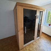 Delta SPA AURA 160 Infrarot-Sauna mit Holzrahmen und schwarzer Glastür in hellem Wohnzimmer