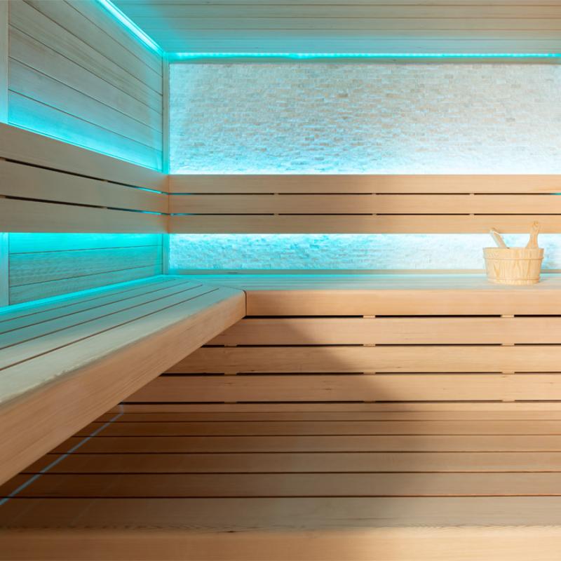 Moderne Infrarot-Sauna mit Hemlock-Holzausstattung, türkiser LED-Beleuchtung an Wand und Decke, Holzbänken und Saunaeimer auf der Ruhebank, Innenansicht