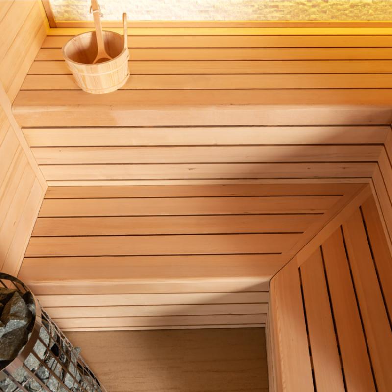 Innenansicht einer Holzsauna mit hellen Hemlock-Holzbänken, Holzdecke mit Beleuchtung, hölzernem Saunaeimer mit Kelle und Saunakelle im Hintergrund
