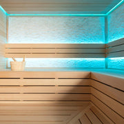 Moderne Infrarot-Sauna mit hellem Hemlock-Holz, großen beleuchteten Panoramafenstern mit türkis-blauer LED-Beleuchtung und hölzernem Saunaeimer auf der Rückenlehne