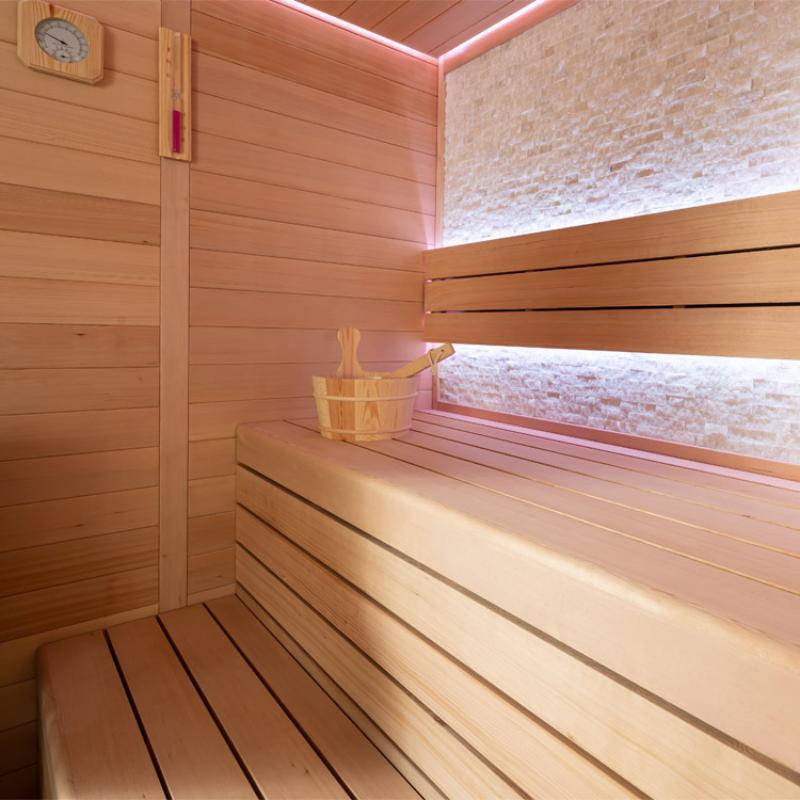 Moderner Saunaraum mit Hemlock-Holzverkleidung, zweistufigen Bänken, Saunalöffel, Sauna-Thermometer und rosa-weißer Beleuchtung an der Decke und hinter den Rückenlehnen