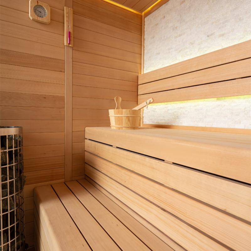 Modernes Inneres einer Hemlock-Sauna mit hellen Holzbänken, Thermometer an der Wand, Saunakübel mit Kelle und Saunaofen links sichtbar.