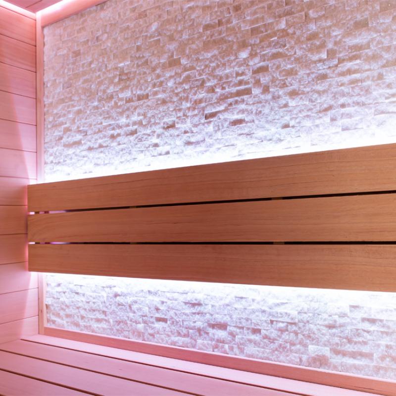 Moderne Infrarot-Sauna mit Hemlock-Holzbank, beleuchtet durch rosafarbene LED-Strahler und strukturierte Salzsteinwand im Innenraum