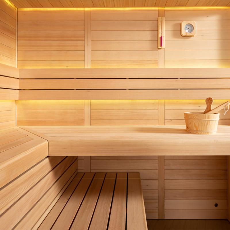 Modernes Saunainneres aus hellem Hemlock-Holz mit zwei Sitzbänken übereinander, Thermometer, Holzeimer mit Kelle und warmer LED-Beleuchtung