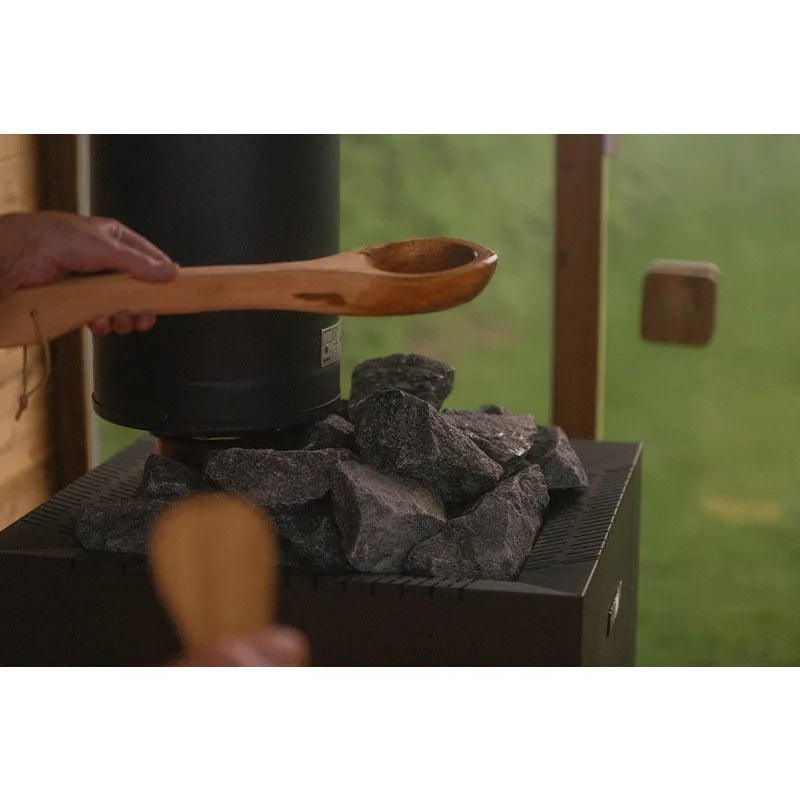 Hekla Cube Gartensauna mit Saunaofen und Aufgusssteinen, Person schöpft Wasser mit Holzkelle