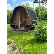 Herkus Large Outdoor-Gartensauna in dunkelbrauner Holzoptik mit schwarzem Schindeldach und integriertem Vorraum, aufgestellt im gepflegten Garten mit Rasen und Nadelbaum.