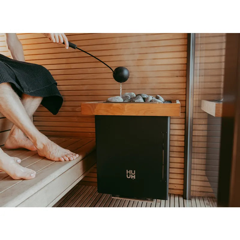 Huum Core Wall Combi Wandofen mit Saunasteinen in moderner Sauna, Person gießt Wasser auf Steine