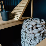 HUUM HIVE Mini Saunaofen mit Saunasteinen in Metallkorb, Sauna im Hintergrund, moderne minimalistische Wellness-Szenerie