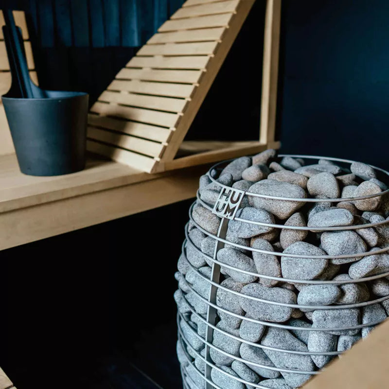 HUUM HIVE Mini Saunaofen mit Saunasteinen in Metallkorb, Sauna im Hintergrund, moderne minimalistische Wellness-Szenerie