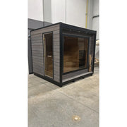 KOBA Cube Gartensauna - Moderne quadratische Form mit schwarzem Rahmen, grauem Holzaußenbau und großem Glasfenster, Innenraum mit Holzbänken sichtbar