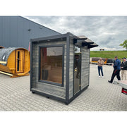 KOBA Cube Gartensauna - Moderne quadratische Form mit grauer Holzverkleidung und großem Fenster auf Ausstellungsgelände