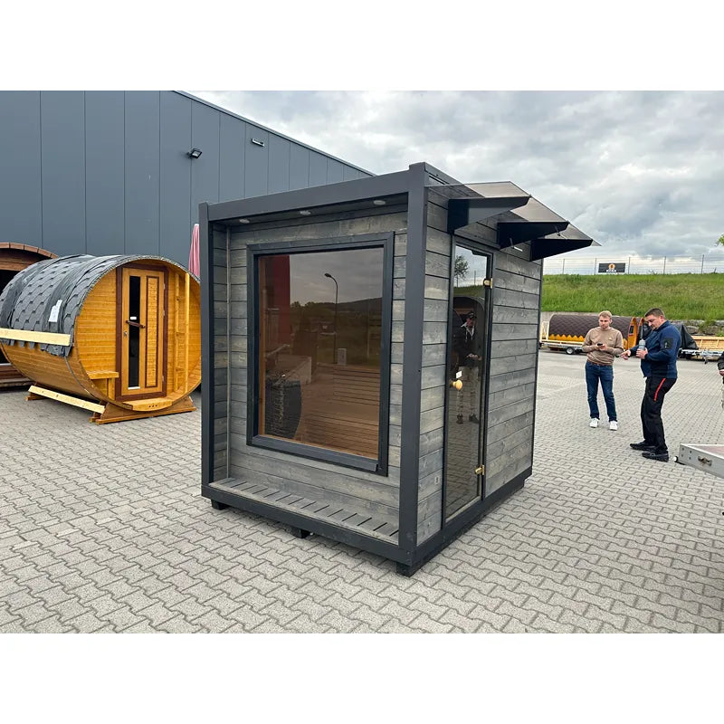 KOBA Cube Gartensauna - Moderne quadratische Form mit anthrazitgrauer Holzverkleidung und großem Fenster auf Ausstellungsgelände