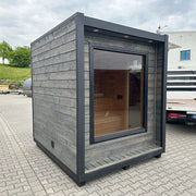 KOBA Cube Gartensauna mit moderner quadratischer Form, grauem Holzrahmen und großem Glaselement auf gepflastertem Untergrund