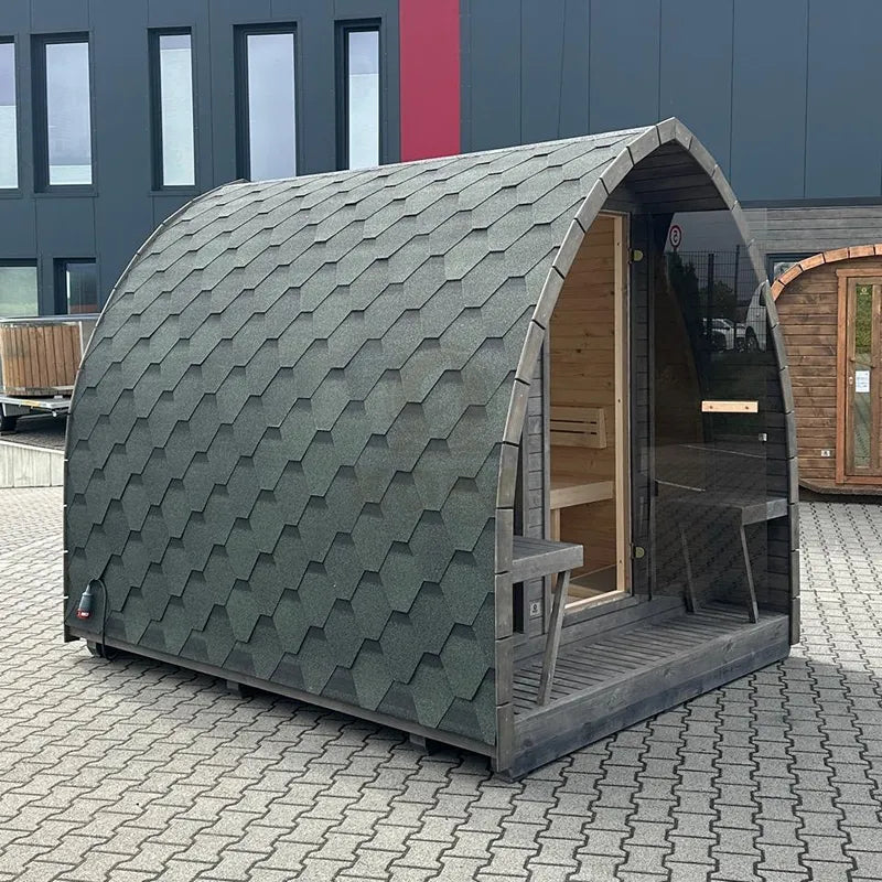 KOBA Iglu Gartensauna mit charakteristischer Iglu-Form, grünes Schindeldach, Holzverkleidung, auf Pflasterplatz vor modernem Gebäude