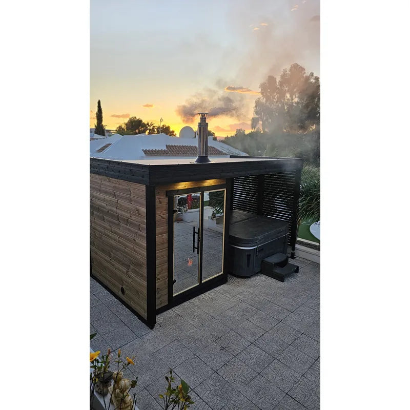 Leil Patio S Plus Gartensauna mit Überdachung im Garten bei Sonnenuntergang, schwarze Holzkonstruktion mit Glastüren und Kaminschornstein