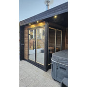 Leil Patio S Plus Gartensauna mit Überdachung im Garten mit beleuchteter Terrasse und Glastüren