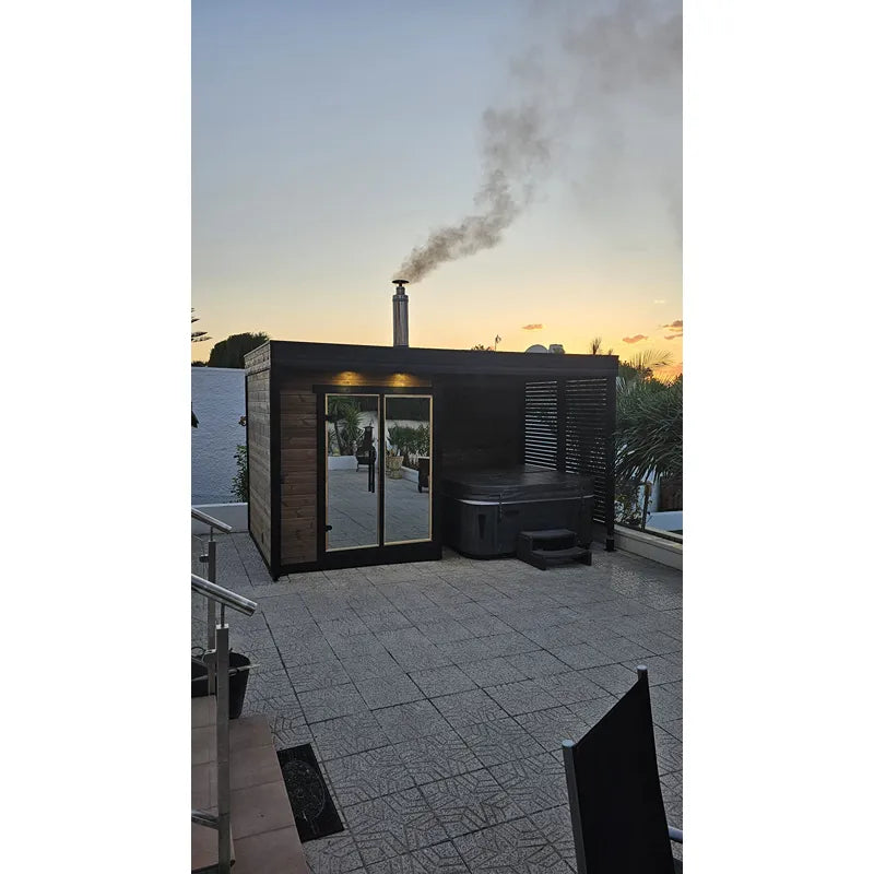 Leil Patio S Plus Gartensauna mit Überdachung von Leil Saunas in schwarzem Design auf Terrasse bei Sonnenuntergang, Rauchentwicklung aus Kamin sichtbar